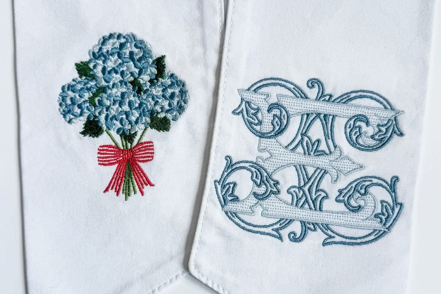 White fabric wreath sash with pointed ends, featuring embroidered details: one with a blue hydrangea bouquet and red ribbon, and the other with a blue monogram design.