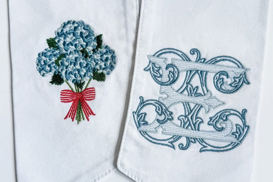 White fabric wreath sash with pointed ends, featuring embroidered details: one with a blue hydrangea bouquet and red ribbon, and the other with a blue monogram design.